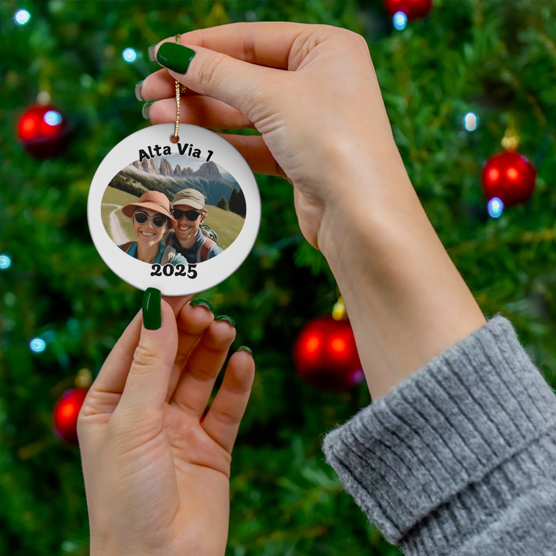 Person holding a personalized Alta Via 1 ornament with custom photo and year 2025 in front of a decorated Christmas tree, highlighting the option to upload your own hiking picture and showcasing the size of the ornament.