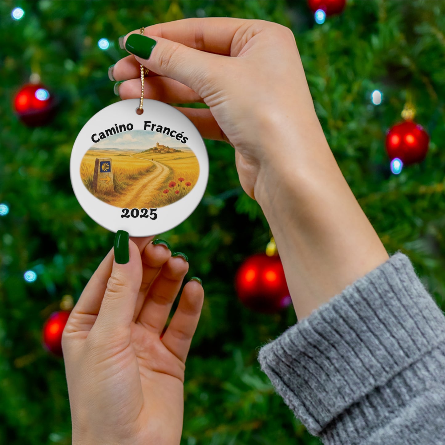 Hand holding Camino Francés round ceramic ornament in front of a decorated Christmas tree. Meseta watercolor trail scene with Camino way marker and year 2025. Ideal Camino pilgrimage keepsake.