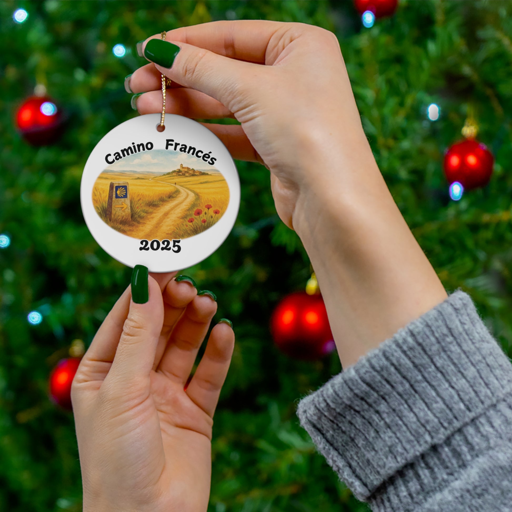 Hand holding Camino Francés round ceramic ornament in front of a decorated Christmas tree. Meseta watercolor trail scene with Camino way marker and year 2025. Ideal Camino pilgrimage keepsake.