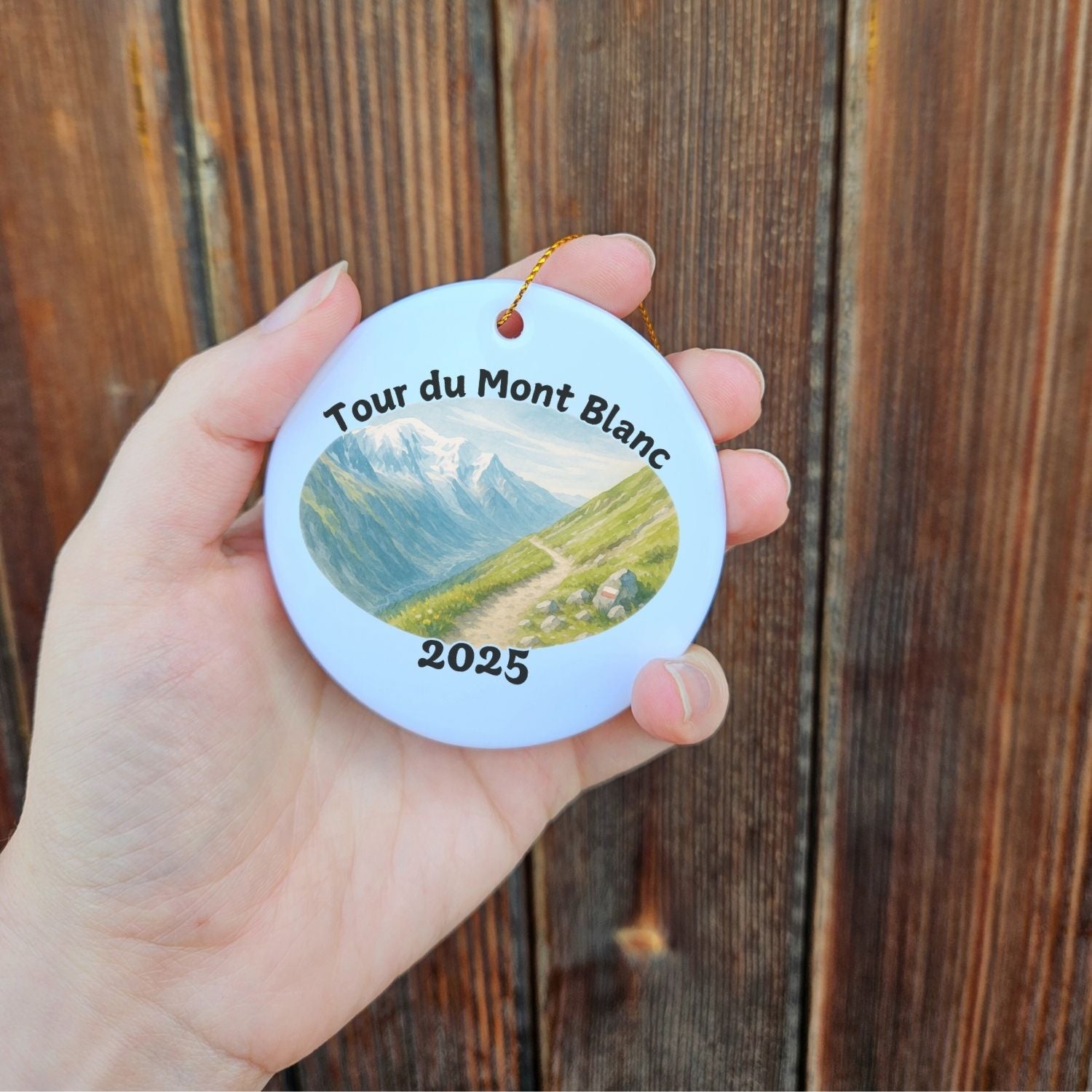 Tour du Mont Blanc hiking keepsake ornament held in hand, showing 2025 watercolor mountain trail artwork with gold cord.