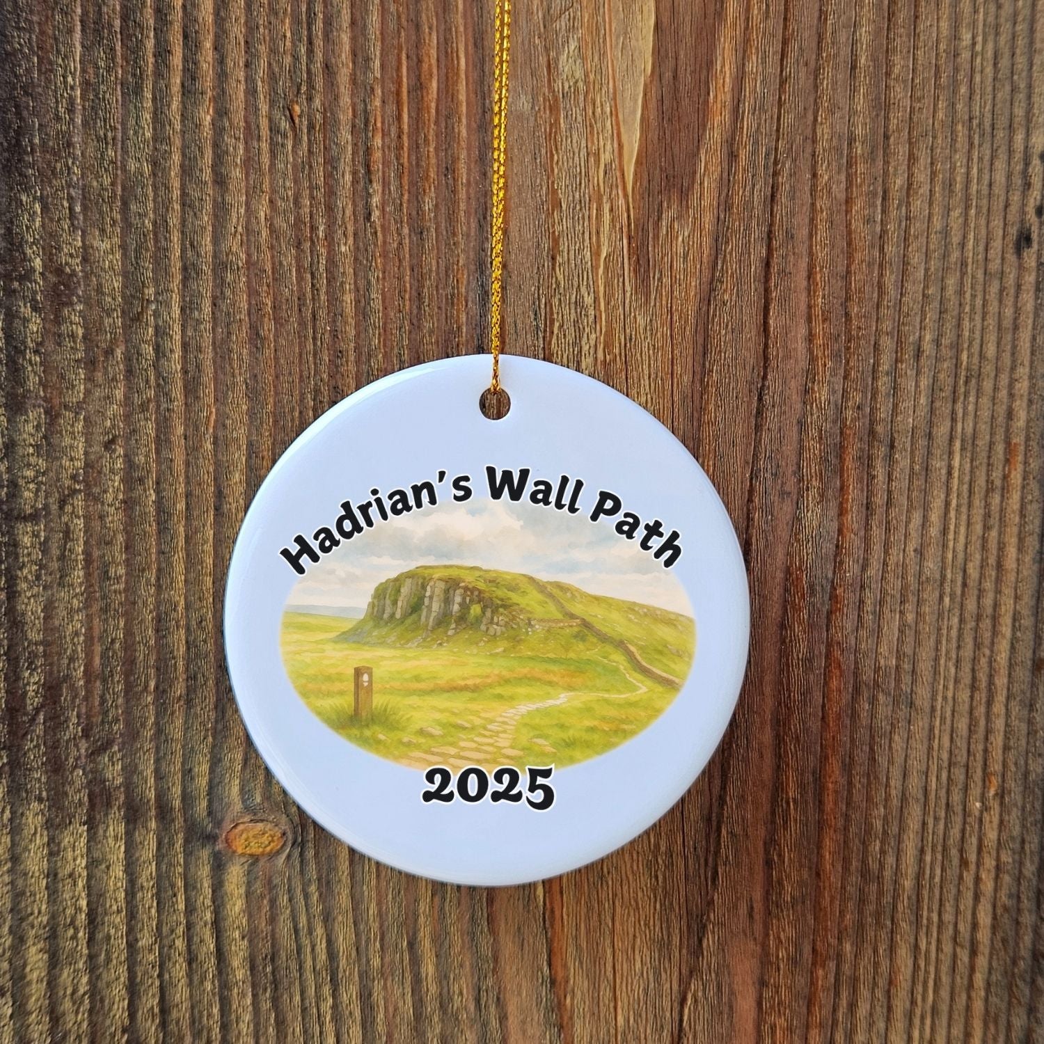 Close-up of the Hadrian’s Wall Path ornament with Steel Rigg landscape design, hanging on a wooden backdrop with golden cord.