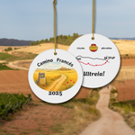 Camino Francés ceramic keepsake ornament with watercolor front and stylized map back, hanging on gold cord, shown against the Camino trail. Perfect gift for pilgrims, year customizable.