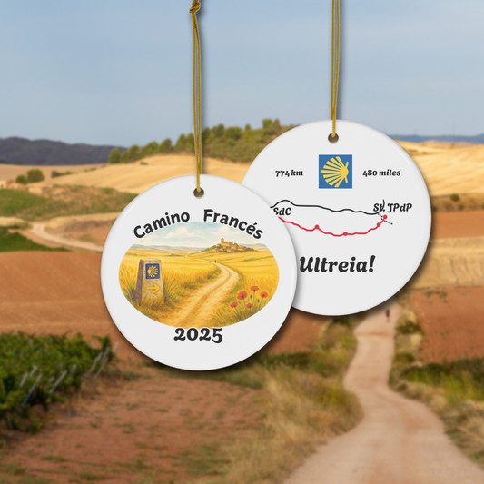 Camino Francés ceramic keepsake ornament with watercolor front and stylized map back, hanging on gold cord, shown against the Camino trail. Perfect gift for pilgrims, year customizable.